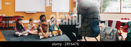 Man teaching an elementary school class using a digital tablet. Teacher is sitting in front of his students and is reading a story from an ebook. Educ Stock Photo
