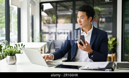 Smart and focused millennial Asian businessman or executive manager holding his smartphone and using laptop computer at his desk in the office. Stock Photo