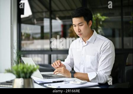 Focused and smart millennial Asian businessman reading business online documents on his laptop at his desk in the office. Stock Photo