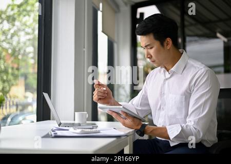 Concentrated and professional millennial Asian businessman or male manager managing his work on digital tablet, remote working at a cafe. Stock Photo