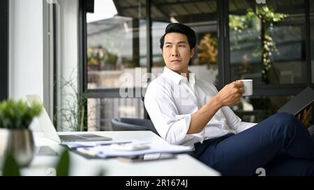 Successful and inspired millennial Asian businessman or male CEO looking out the window, thinking and planning his business while having his morning c Stock Photo