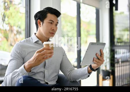 Handsome millennial Asian businessman having morning coffee and using his tablet in an office lounge. Stock Photo