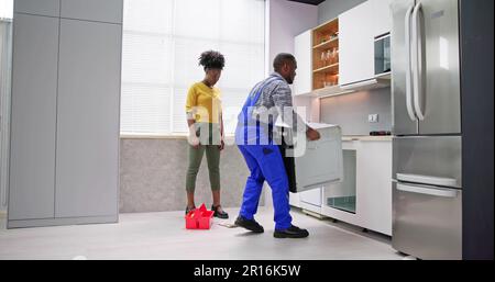 Young Woman Looking At Male Worker In Overall Repairing Oven In Kitchen Stock Photo