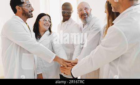 A diverse team of three men and three women, wearing lab coats and safety glasses, join hands in a symbol of teamwork, collaboration, and success in a Stock Photo