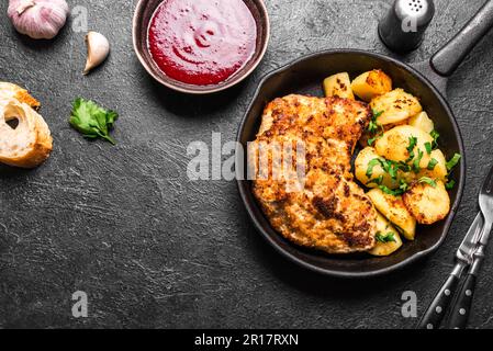 German schnitzel with roasted potatoes and cranberry sauce on  black cast iron pan, copy space. Homemade breaded pork meat chop. Wiener Schnitzel. Stock Photo