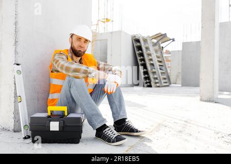 Builder repairman, foreman in safety helmet and vest sitting down to rest at workplace in building next to tool box and ruler. Stock Photo