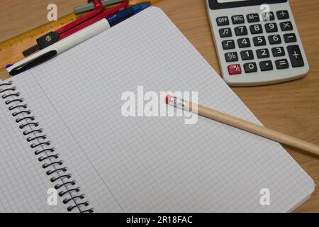 Top view of a student wooden table with an open notebook with grid sheets, a pencil with an eraser, a calculator, a compass and a ruler Stock Photo