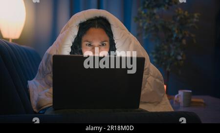 Hispanic freelancer girl Latino woman Indian Arabian female at night evening home at couch sofa under blanket cover duvet working with laptop watching Stock Photo