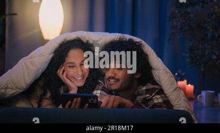 Multiracial couple man and woman at night home using mobile phone shocked amazed with online news shock wonder smartphone social media at evening on Stock Photo