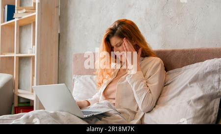 Freelancer Caucasian sick woman working remote from home bed bad feeling headache. Ill student girl studying distant with laptop in bedroom suffer Stock Photo