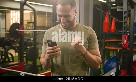 Excited happy African American trainer gym instructor guy sportsman male athlete bodybuilder man in sport club champion winner with mobile phone Stock Photo
