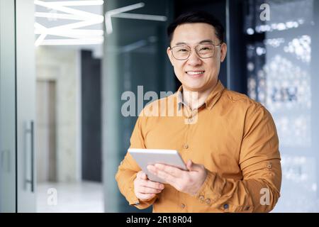 Portrait of young programmer with tablet computer inside office,asian man smiling and looking at camera, man at workplace testing online applications standing near window. Stock Photo