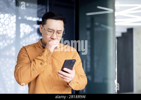 Asian businessman inside office building upset and sad reading bad news on mobile phone, business owner investor using smartphone standing near window. Stock Photo