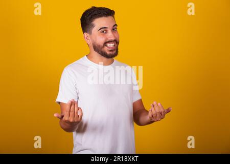 Brazilian Latin American man, with gesture calling, come, come, inviting, receiving, welcoming, happy, smiling, autruism, yellow background, space for Stock Photo