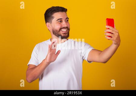 Brazilian Latin American man, Taking selfie with cell phone, on video call, talking, smiling, photographing. Yellow background. Stock Photo