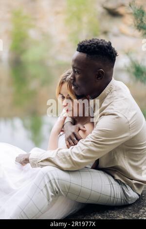 Happy interracial couple newlyweds sits on rock and embraces against beautiful background of lake. Closeup. Concept of love relationships and unity be Stock Photo