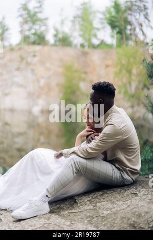 Happy interracial couple newlyweds sits on rock and embraces against beautiful background of lake, forest and canyon. Concept of love relationships an Stock Photo