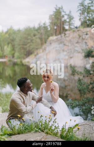 Happy interracial couple newlyweds sits and has a fun near dandelions against background of lake, forest and canyon. Concept of love relationships and Stock Photo