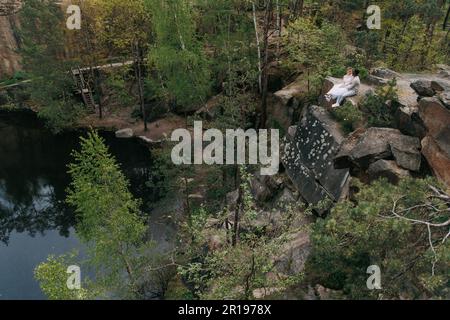 Young interracial couple newlyweds sits on rock and talks against background of forest, lake and canyon. Concept of love relationships and unity betwe Stock Photo