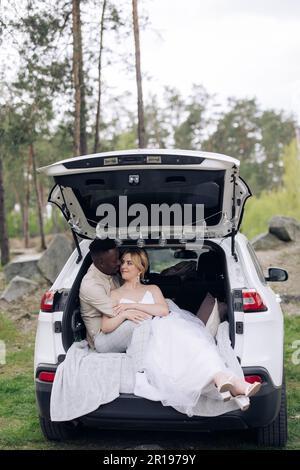 Happy interracial couple newlyweds sits in opened car trunk and embraces against background of forest. Concept of love relationships and unity between Stock Photo