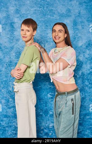 Smiling tattooed and long haired gay man hugging queer friend crossing arms during lgbt pride moth celebration on textured blue background Stock Photo
