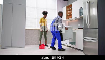 Young Woman Looking At Male Worker In Overall Repairing Oven In Kitchen Stock Photo