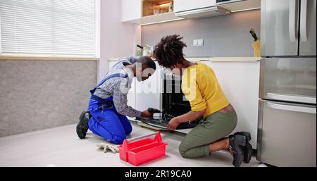 Male Worker Repairing Oven Appliance In Presence Of Young Woman In Kitchen Room Stock Photo