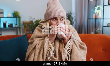 Sick senior grandmother woman in plaid shivering from cold on sofa drinking hot tea in unheated apartment without heating due debt. Unhealthy ill mature pensioner feeling discomfort try to warming up Stock Photo