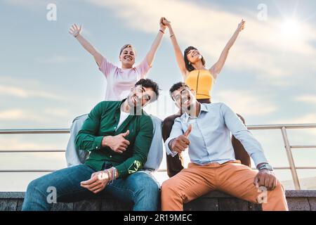 A group of four multicultural friends at the waterfront. The two men sit on a bench, gesturing approval, while the woman and non-binary person raise t Stock Photo