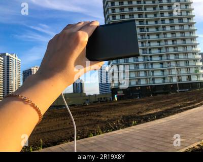 girl holding a charging block on the background of a multi-storey residential building. she is wearing a gold fashion bracelet. the building is made i Stock Photo