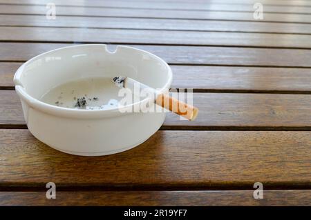 A white ceramic ashtray with a cigarette and ash with a black lighter for smoking stands on a wooden table in a cafe. Stock Photo