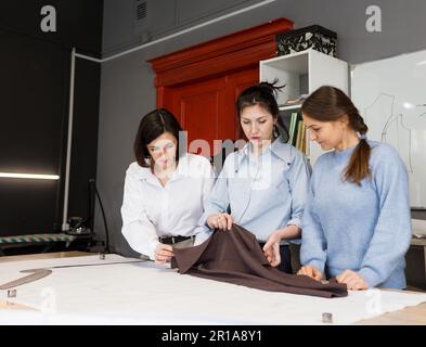 Three female fashion designers work together in a tailoring studio. A group of women dressmakers discusses fabric, pattern and sketch in a sewing work Stock Photo