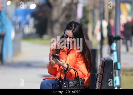 17 March 2025, Istanbul, Turkey: Young woman in bright orange jacket scrolling smartphone while sitting on a park bench on a sunny early-spring day Stock Photo