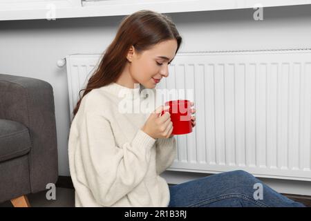 Woman holding cup with hot drink near heating radiator indoors Stock Photo