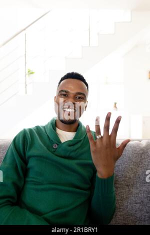 Portrait of african american young man smiling and showing wedding ring while sitting on sofa Stock Photo