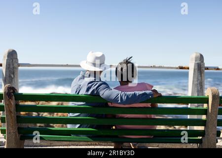 Senior african american couple sitting on bench and embracing on promenade at seaside Stock Photo