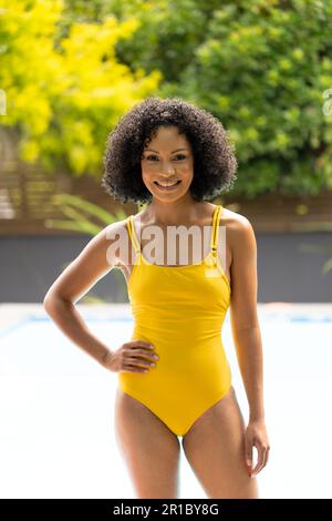 Portrait of happy african american woman in swimsuit standing with hand on hip by pool in garden Stock Photo