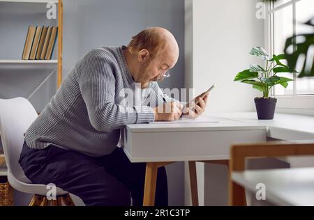 Senior man sitting at table at home, looking at his cell phone, and taking notes Stock Photo