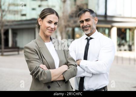Confident and smiling business people looking at camera in the street with their arms crossed. Young businesswoman with a senior businessman behind Stock Photo