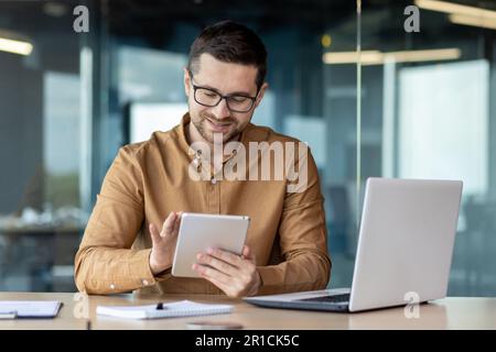 Portrait of a smiling young man designer, engineer, architect sitting in the office behind a notebook and works, draws, doodles online on the tablet he is holding in his hands. Stock Photo