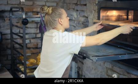 A female baker inserts a baking sheet into the oven in a bakery Stock Photo
