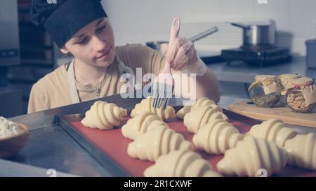 baker girl covers croissants with egg yolk. Stock Photo