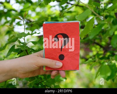 A woman holding a stack of colorful paper notes with question marks on them in her hand against a background of foliage. Close up. Stock Photo