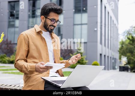 Young Hindu student studying sitting on bench outside university campus, man using laptop for video call, taking notes with pen. Stock Photo