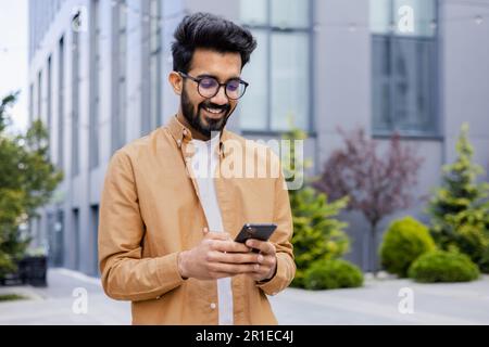 Successful Indian young businessman outside office building walking in daytime, man holding phone in hands, businessman dialing, browsing online pages, programmer engineer in glasses. Stock Photo