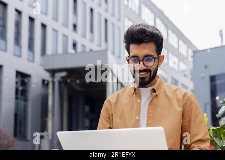 Young successful businessman working with laptop remotely sitting on bench outside office building, Hindu programmer in shirt smiling happy with achievement results and success. Stock Photo