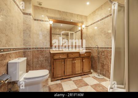 A small bathroom with a white shower stall, a wooden cabinet with a marble top, a checkered floor, tiled borders on the walls, and a long mirror with Stock Photo