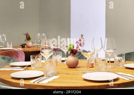 Round wooden dining tables of a restaurant with glassware next to the dishes and some decorative plants Stock Photo