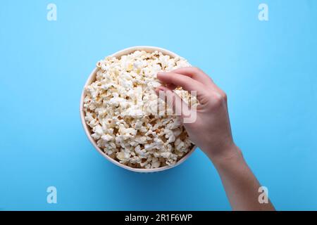 Woman taking delicious popcorn from paper bucket on light blue background, top view Stock Photo