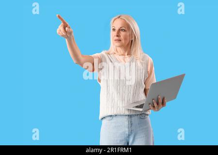 Mature female programmer with laptop pointing at something on blue background Stock Photo